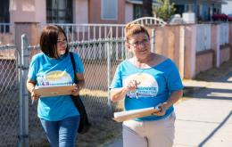 Two Latina women wearing bright blue t-shirts with a health coalition logo on them walk through a residential neighborhood with clipboards.  